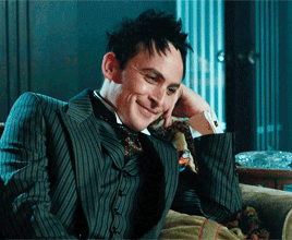 Oswald Cobblepot portrayed by Robin Lord Taylor in the TV show "Gotham", from a season 4 episode. He is wearing a suit and bowtie, a purple streak is in his hair and he is smiling. There is a candle holding chandelier behind him.